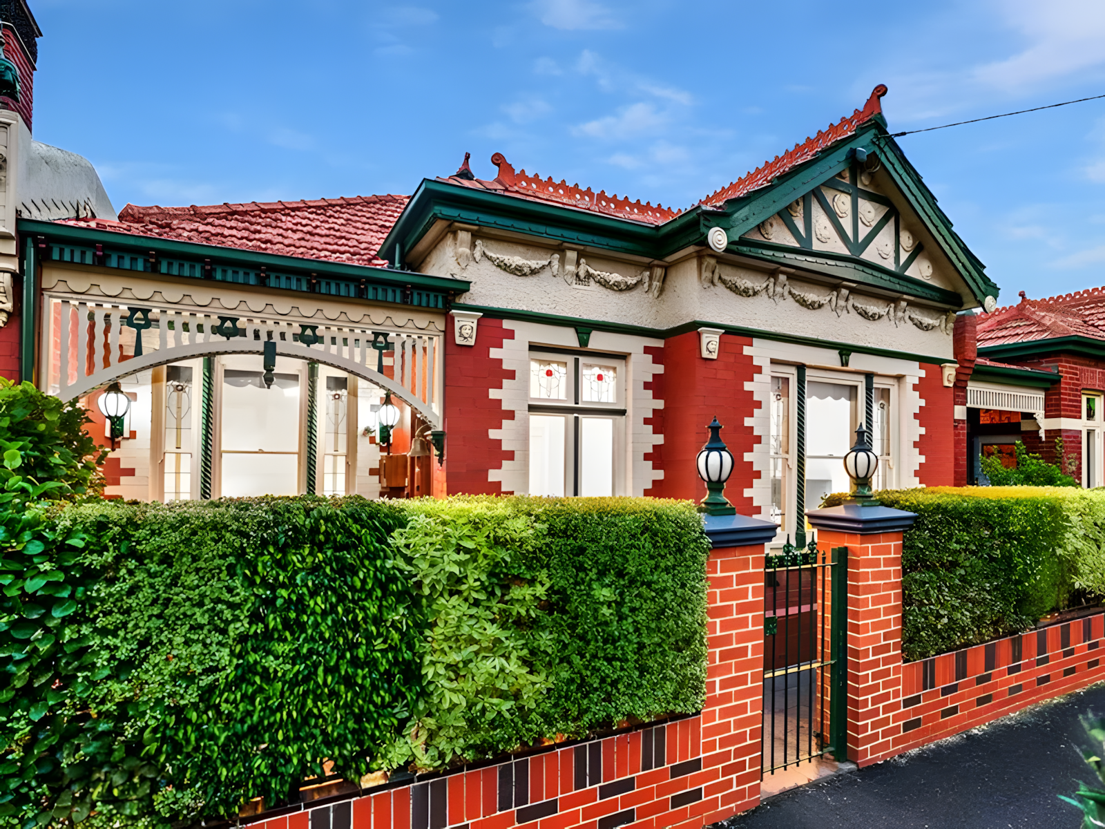 A charming, freshly painted heritage house with red brick and ornate detailing. It features large windows, a green and red tiled roof, and a decorative fence with lanterns.